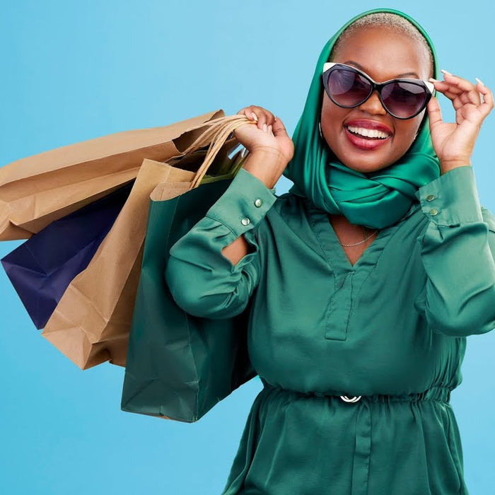 A smiling woman in a green dress and sunglasses holding several shopping bags.