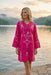 A woman wearing a bright pink embroidered tunic dress stands by a calm lake at sunset, with mountains in the background.