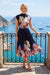 A woman stands on a stone terrace overlooking a blue ocean and rocky coastline, wearing a dark navy floral pleated skirt from Frank Lyman