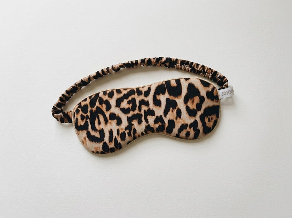 A leopard print sleep mask with an elastic strap, shown on a plain white background. The brand name 'JOHN' is visible on a small fabric tag at the end of the strap.