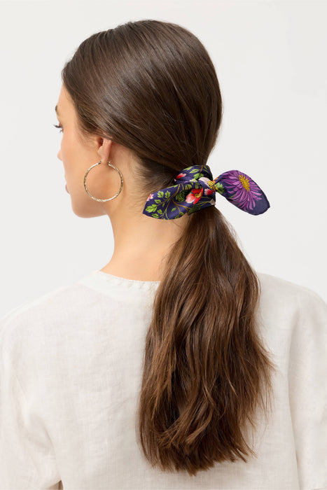 A woman shown from the back wearing a dark brown hair ponytail secured with a colorful floral silk scrunchie. The scrunchie features purple, green, and pink floral patterns on a dark background.