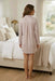 A woman stands barefoot in a bedroom, viewed from the back, wearing a light blush pink long-sleeve button-up shirt dress with a subtle textured pattern. The bedroom features a neutral beige bed, white linens, and a window with sheer curtains.