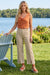 A woman stands on a grassy lawn by a lake wearing a sleeveless rust-orange cowl-neck top and beige cropped wide-leg pants with side slits from Frank Lyman