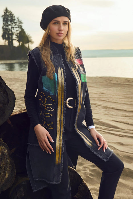 A woman posing on a sandy beach wearing a dark denim longline vest with colorful patchwork panels, gold embroidery, and fringe details, paired with a black beret, black top, and black pants.