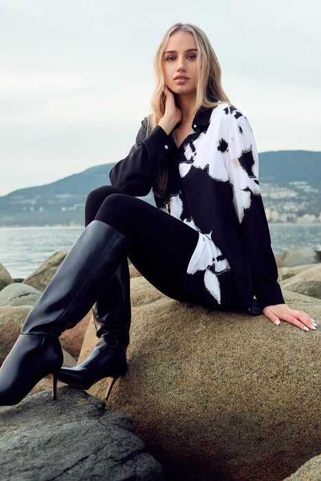 A woman sits on large rocks by the water wearing a black and white patterned long-sleeve top, black pants, and black knee-high heeled boots.