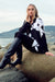 A woman sits on large rocks by the water wearing a black and white patterned long-sleeve top, black pants, and black knee-high heeled boots.
