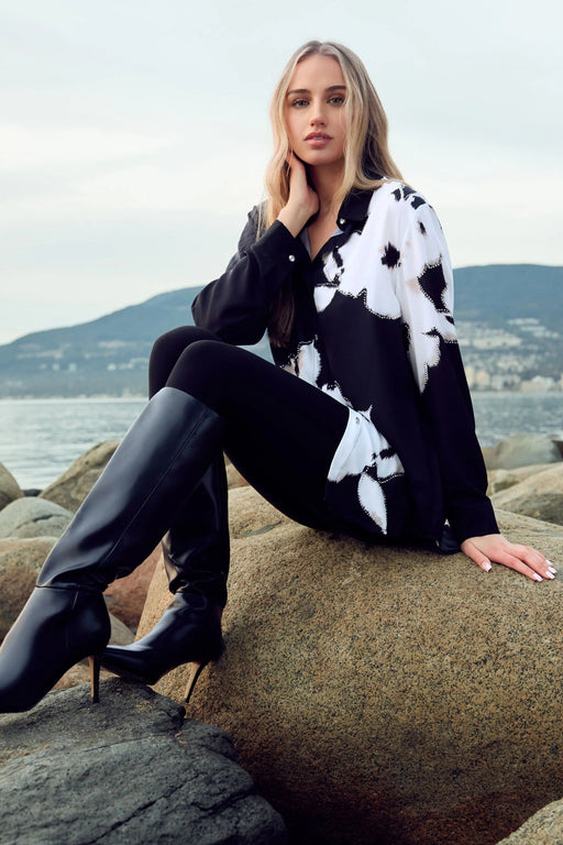 A woman sits on large rocks by the water wearing a black and white patterned long-sleeve top, black pants, and black knee-high heeled boots.