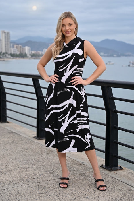 A woman stands outdoors by a waterfront railing wearing a sleeveless black midi dress with a bold white abstract brushstroke pattern from Frank Lyman