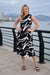 A woman stands outdoors by a waterfront railing wearing a sleeveless black midi dress with a bold white abstract brushstroke pattern from Frank Lyman