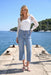A woman stands on a concrete dock by the sea wearing a white scoop-neck top with sheer lace sleeves and Frank Lyman light-wash blue cropped jeans with button details at the hips, paired with white heeled sandals.
