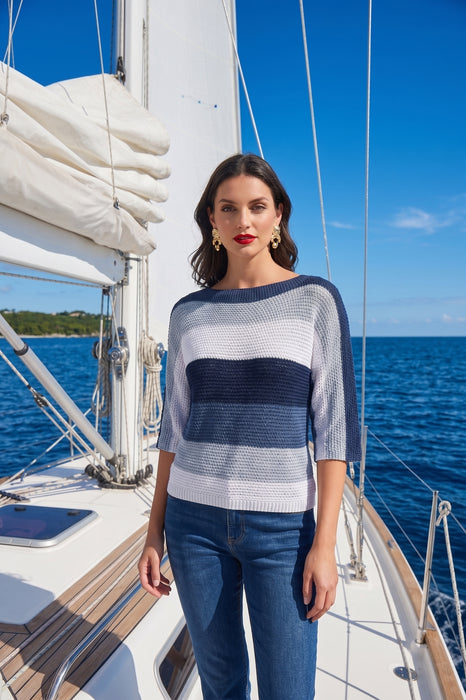 A woman stands on a sailboat deck wearing a blue and white striped knit sweater from Frank Lyman and blue jeans, with the blue ocean and sky in the background.