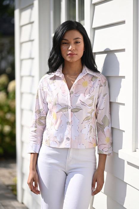 A woman stands outdoors against a white house wall wearing a light pink button-up jacket with a soft floral print from Frank Lyman