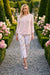 A woman walks down a gravel path in a formal garden wearing a light pink cardigan and white cropped pants with a delicate floral print from Frank Lyman