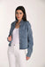 A woman modeling a cropped, long-sleeve blue denim jacket with a subtle textured pattern, worn open over a white tank top and white pants, against a plain light background.