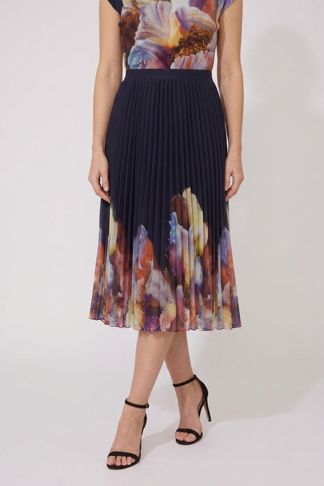 A model wearing a pleated midi skirt with a dark navy base and a colorful abstract floral print at the waist and hem, paired with black strappy high-heeled sandals against a plain light gray studio background.