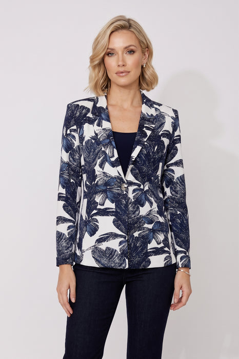 A woman modeling a navy blue and white tropical leaf print blazer over a black top and dark jeans, standing against a light gray studio background.