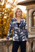 A woman stands outdoors in front of a stone balustrade and building facade, wearing a navy blue and white tropical floral print blazer from Frank Lyman.