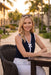 A woman sitting at an outdoor patio table wearing a navy sleeveless top with a white bow detail and contrast trim from Frank Lyman