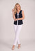 A woman modeling a sleeveless navy blue top with white trim and a white bow at the neckline, paired with white pants and beige heels, against a light gray studio background.