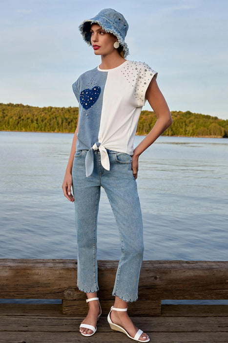 A woman stands on a wooden dock by a calm lake wearing a light blue denim bucket hat, a white and blue tie-front top with a blue heart patch, light-wash cropped jeans, and white wedge sandals.