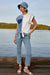 A woman stands on a wooden dock by a calm lake wearing a light blue denim bucket hat, a white and blue tie-front top with a blue heart patch, light-wash cropped jeans, and white wedge sandals.