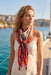 A woman on a boat wearing a cream camisole top and a vibrant red, orange, and black paisley patterned scarf tied around her neck, with blue water and cliffs in the background.