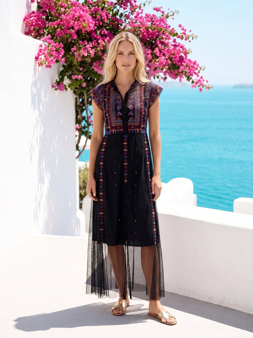 A woman modeling a black midi dress with colorful bohemian embroidery and sheer tulle hem, standing outdoors on a white terrace with a blue sea and pink bougainvillea flowers in the background.