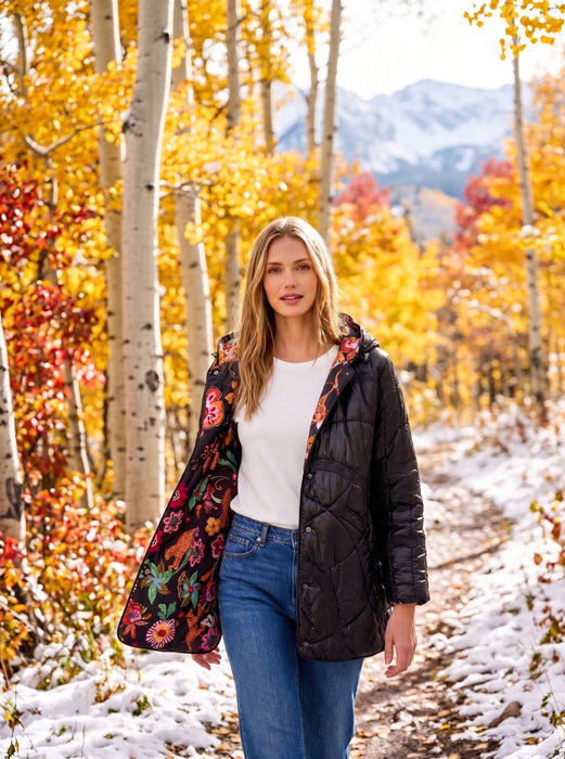 A woman stands on a snowy path surrounded by bright autumn trees, wearing a black quilted puffer jacket with a colorful floral lining over a white top and blue jeans.