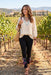 A woman walking through a sunny vineyard wearing a cream lace-trim blouse, black pants with colorful floral embroidery near the cuffs, layered turquoise necklaces, and carrying a woven straw tote bag.