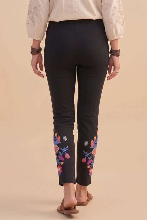 A person shown from the back wearing black cropped pants with colorful floral embroidery near the lower calves, paired with a cream blouse and brown sandals, against a plain beige studio background.