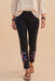 A model is shown from the waist down wearing black cropped leggings with colorful floral embroidery near the lower calves, paired with a cream blouse and brown sandals, against a solid beige background.