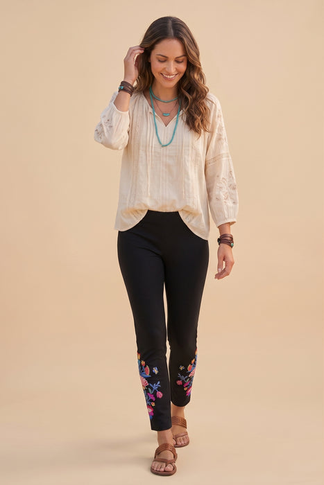 A woman modeling a cream-colored embroidered blouse with black pants featuring colorful floral embroidery near the cuffs, styled with layered turquoise necklaces and brown sandals, against a solid beige background.