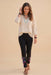 A woman modeling a cream-colored embroidered blouse with black pants featuring colorful floral embroidery near the cuffs, styled with layered turquoise necklaces and brown sandals, against a solid beige background.