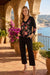 A woman stands on a sunlit stone terrace with arches and a coastal view in the background, wearing a black long-sleeve top with colorful floral embroidery and black cropped pants.
