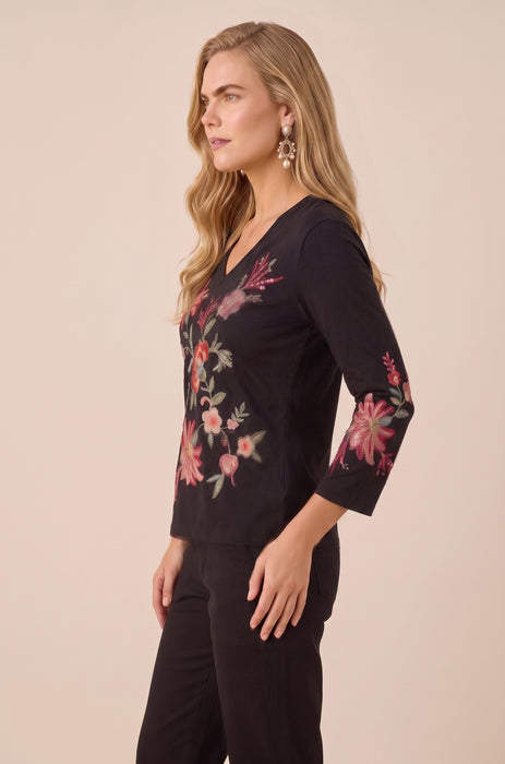 A woman modeling a black three-quarter sleeve top with pink and green floral embroidery, paired with black pants, against a solid beige background.