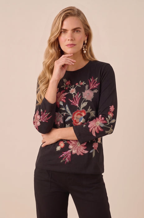 A woman modeling a black long-sleeve top with vibrant pink, red, and green floral embroidery, paired with black pants, against a solid beige background.