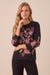 A woman modeling a black long-sleeve top with vibrant pink, red, and green floral embroidery, paired with black pants, against a solid beige background.