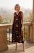 A woman stands on a stone balcony wearing a sleeveless, V-neck midi dress with intricate floral and butterfly embroidery in dark brown, burgundy, and gold tones. The background shows a softly blurred vineyard landscape.