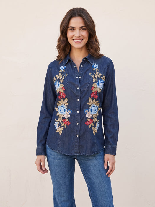 A woman wearing a dark blue denim button-up shirt with colorful floral embroidery on the front, paired with blue jeans, standing against a plain beige background.