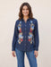 A woman wearing a dark blue denim button-up shirt with colorful floral embroidery on the front, paired with blue jeans, standing against a plain beige background.