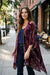 A woman stands on a city sidewalk wearing a long, open-front purple and burgundy patterned cardigan over a navy top and dark jeans. Trees, buildings, and outdoor cafe seating are visible in the background.