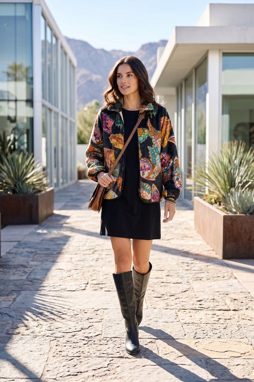 A woman walking outdoors on a stone pathway between modern glass buildings, wearing a colorful floral-patterned jacket over a black dress and dark knee-high boots, with a brown shoulder bag.