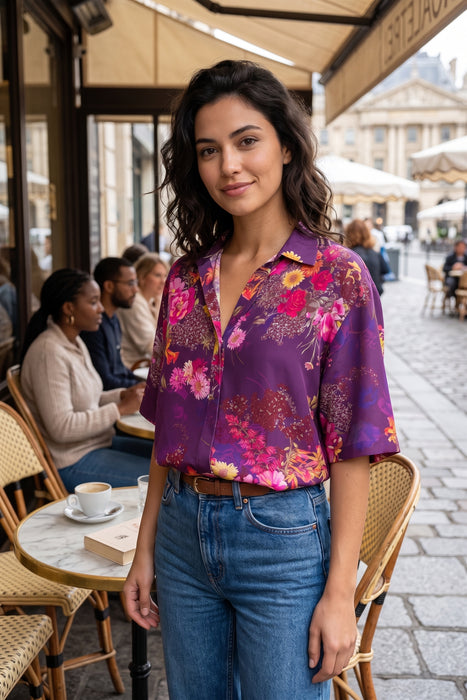 A woman stands at a Parisian-style cafe wearing a purple short-sleeve button-up blouse with a vibrant pink, red, and yellow floral print, paired with blue jeans.