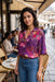 A woman stands at a Parisian-style cafe wearing a purple short-sleeve button-up blouse with a vibrant pink, red, and yellow floral print, paired with blue jeans.