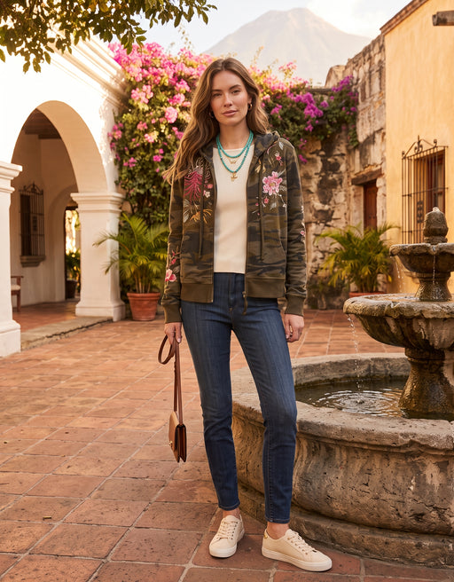 A woman stands in a sunlit courtyard wearing a camouflage bomber jacket with floral embroidery on the sleeves and chest, a cream knit top, blue skinny jeans, and white sneakers. A stone fountain, archway, and flowering plants are visible in the background.