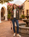 A woman stands in a sunlit courtyard wearing a camouflage bomber jacket with floral embroidery on the sleeves and chest, a cream knit top, blue skinny jeans, and white sneakers. A stone fountain, archway, and flowering plants are visible in the background.