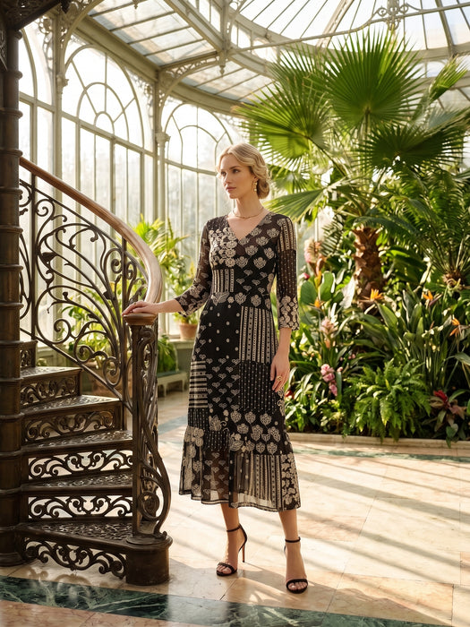 A woman models a black midi dress with beige and white geometric patterns in a sunlit greenhouse with an ornate spiral staircase.