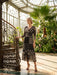 A woman models a black midi dress with beige and white geometric patterns in a sunlit greenhouse with an ornate spiral staircase.