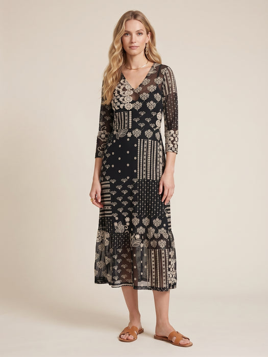 A woman modeling a black midi dress with a beige and white patchwork-style bohemian print, featuring sheer three-quarter sleeves and a V-neckline, set against a plain beige studio background.