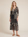 A woman modeling a black midi dress with a beige and white patchwork-style bohemian print, featuring sheer three-quarter sleeves and a V-neckline, set against a plain beige studio background.